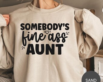 Puede incluir: Una sudadera de color arena con el texto negro "SOMEBODY'S fine ass AUNT". El diseño incluye un corazón y líneas decorativas. La sudadera tiene cuello redondo y mangas largas.