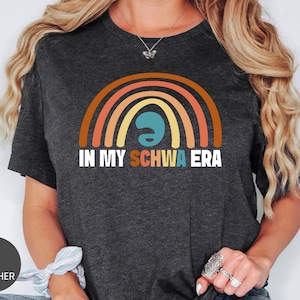 May include: Dark grey heather t-shirt featuring a colorful rainbow design with the text "IN MY SCHWA ERA". The rainbow includes shades of orange, yellow, and teal. The shirt is a casual wear item.