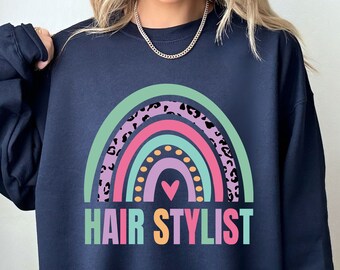 May include: A navy blue sweatshirt with a rainbow graphic and the text "HAIR STYLIST". The rainbow is made up of pink, purple, green, and yellow stripes with a black leopard print pattern on the outer edges. A small pink heart is in the center of the rainbow.