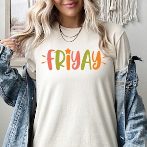 May include: A woman wearing a light beige t-shirt with the word "FRIYAY" written in colorful letters. The shirt has a star above the word "FRIYAY". She is wearing a denim jacket over the shirt.