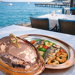 Puede incluir: Un plato de servicio de cobre con textura martillada y asa dorada, parcialmente abierto para revelar pescado a la parrilla, camarones, tomates y rodajas de limón. El plato está sobre un mantel blanco cerca del mar.