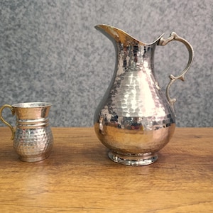 Peut inclure: Une cruche en métal argenté martelé et une tasse assortie plus petite avec une poignée en laiton. Les deux sont sur une surface en bois.