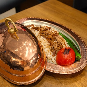Op de afbeelding: Een koperen serveerschaal met een decoratieve handgreep en een bijpassende serveerschaal. De schaal bevat een portie rijst, vlees, een geroosterde tomaat en een groene paprika. De schaal staat op een houten tafel.