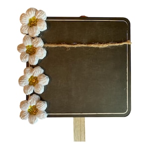 May include: A small wooden sign with a chalkboard surface and a white wooden stake. The sign is decorated with white crocheted flowers with yellow centers. A piece of twine is tied around the sign.