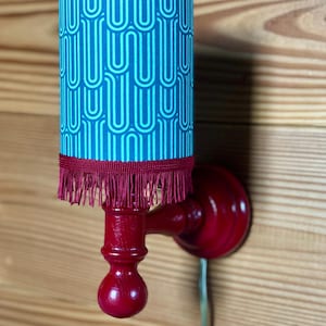 May include: A red wall sconce with a teal blue and white patterned lampshade. The lampshade has a red fringe trim.