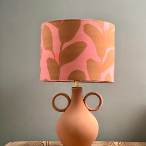 May include: A terracotta table lamp with two circular handles on the base. The lampshade is pink with a brown abstract floral pattern.