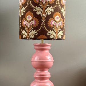 Puede incluir: Lámpara de mesa de estilo vintage con base rosa y pantalla estampada. La pantalla presenta un fondo marrón con diseños florales en naranja, rosa y crema. La lámpara tiene una forma decorativa clásica, adecuada para la decoración del hogar.