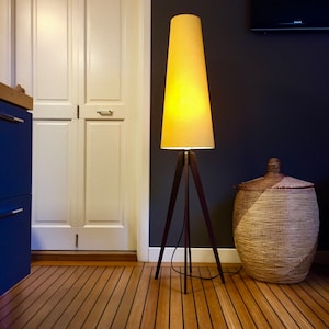 Può includere: Una lampada da terra gialla con una base a treppiede e un paralume conico. La lampada è accesa e illumina la stanza. La lampada è davanti a un muro scuro e un cesto di vimini.