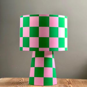 Op de afbeelding: Een roze en groene geruite tafellamp met een conische voet. De lampenkap is cilindrisch en de voet is een kegel.