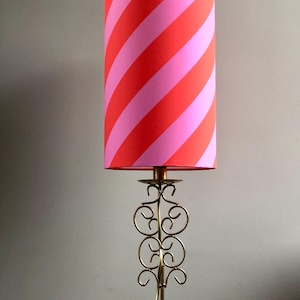 Op de afbeelding: Een tafellamp met een cilindrische lampenkap met diagonale strepen in felrood en lichtroze. De lampvoet is gemaakt van dun, goudkleurig metaaldraad gevormd in decoratieve krulpatronen met een statiefachtige standaard.