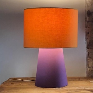 Op de afbeelding: Een tafellamp met een cilindrische oranje stoffen lampenkap en een conische voet die verloopt van een lichtroze tint aan de bovenkant naar een dieppaarse kleur aan de onderkant. De lamp is voorzien van een dun snoer aan de voet.