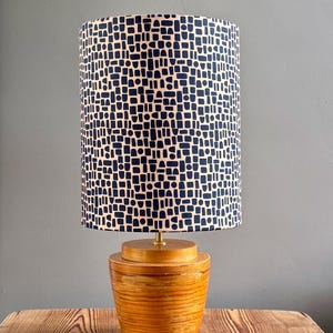 Puede incluir: Una lámpara de mesa con una pantalla cilíndrica que presenta un patrón geométrico azul oscuro sobre un fondo claro. La base está hecha de madera de color marrón claro, redondeada y apilada, con un acabado liso y un pequeño conector de metal dorado.