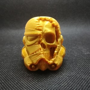 Peut inclure: Une figurine en or imprimée en 3D d'un casque de Stormtrooper de Star Wars avec un motif de crâne. Le casque est fissuré et cassé, révélant le crâne en dessous.