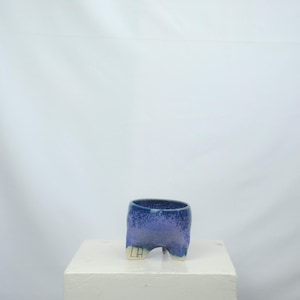 May include: A small, handmade ceramic cup with a unique, three-legged design. The cup features a gradient of blue hues, transitioning from a deep indigo at the rim to a lighter, lavender shade towards the base. The legs are a contrasting off-white color.
