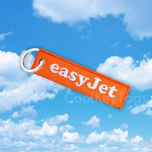Può includere: Portachiavi rettangolare arancione con la scritta "easyJet" in bianco. Ha un anello in metallo argentato. Lo sfondo è un cielo blu con nuvole bianche.