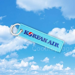 Può includere: Un portachiavi blu chiaro con un cerchio bianco e un contorno rosso, con il testo "KOREAN AIR" in ricamo blu.