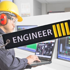 以下が含まれることがあります： 白い文字で「ENGINEER」と書いてある黒いキーホルダーに、黄色のストライプが3本付いている。キーホルダーは、黄色いヘルメットと安全メガネを着用した人が持っている。