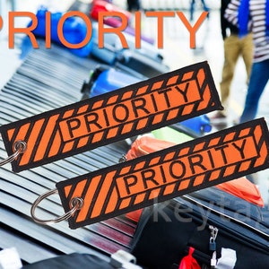 Könnte beinhalten: Zwei orangefarbene und schwarze Gepäckanhänger mit dem Wort "PRIORITY" in schwarzen Buchstaben. Die Anhänger sind an silbernen Schlüsselringen befestigt.