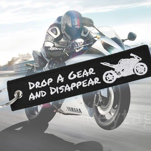 以下が含まれることがあります： 「Drop a gear and disappear」と白く刺繍された黒いオートバイのキーホルダー。キーホルダーには、オートバイの白いシルエットが描かれています。