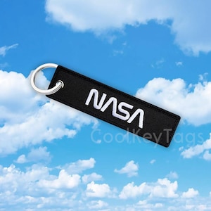 May include: A black key chain with a white NASA logo. The keychain is against a blue sky with white clouds.