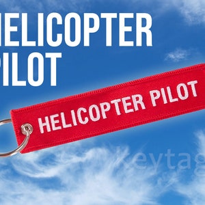 May include: Red fabric keychain with white text that reads "HELICOPTER PILOT". The keychain is against a blue sky with white clouds.