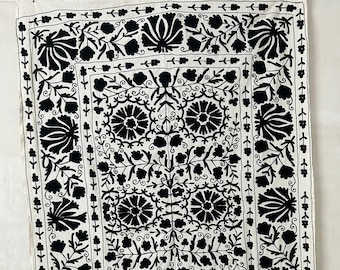 Pode incluir: Uma tapeçaria de parede de tecido branco com um desenho de bordado floral preto. O desenho apresenta um padrão repetitivo de flores e folhas estilizadas.