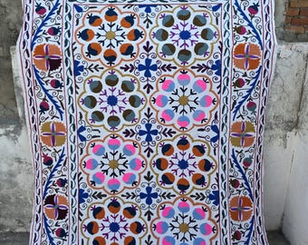 May include: A white textile with a colourful, symmetrical floral pattern. The design features blue, orange, pink, and purple flowers and geometric shapes, framed by a detailed border. The textile is likely a decorative piece, such as a wall hanging or a rug.