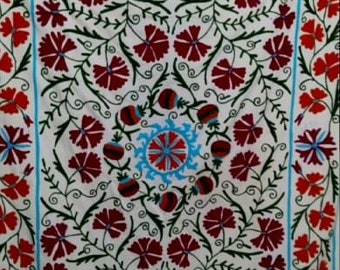 May include: White fabric with a red and blue floral embroidery pattern. The design features a central motif of a stylized flower with a blue center and red petals, surrounded by smaller red flowers and green leaves.