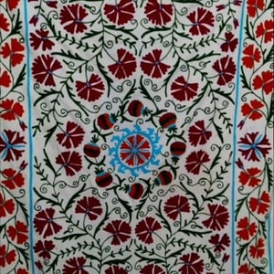 Op de afbeelding: Witte stof met een rood en blauw bloemenborduurpatroon. Het ontwerp heeft een centraal motief van een gestileerde bloem met een blauwe kern en rode bloemblaadjes, omringd door kleinere rode bloemen en groene bladeren.