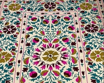 May include: A white fabric with intricate embroidery featuring a floral pattern in shades of pink, teal, brown, and gold. The design includes stylized flowers, leaves, and vines.