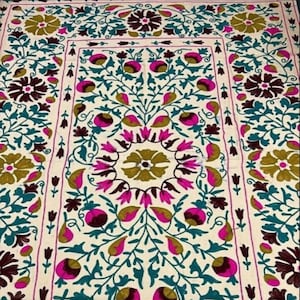 Puede incluir: Una tela blanca con bordado intrincado que presenta un patrón floral en tonos de rosa, azul verdoso, marrón y dorado. El diseño incluye flores, hojas y enredaderas estilizadas.