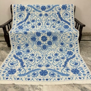 Peut inclure: Textile blanc orné d'un motif floral bleu, drapé sur une chaise en bois foncé. Le motif présente des fleurs et des feuilles stylisées dans différentes nuances de bleu, créant un arrangement symétrique et décoratif. Le textile semble être fait d'une matière tissée douce.