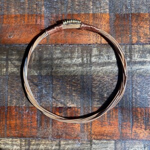 Puede incluir: Un collar circular hecho de múltiples hebras de cuerdas de guitarra de cobre y plata. Las cuerdas están unidas entre sí con pequeñas cuentas doradas.