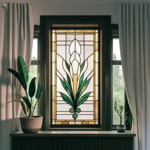 Op de afbeelding: Een glas-in-loodraam met een bloemmotief met een witte bloem en groene bladeren. Het raam is omlijst met donker hout en is geplaatst in een muur met witte gordijnen aan beide kanten.