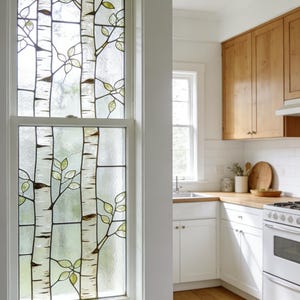 Könnte beinhalten: Buntglasfenster mit Birkenbaum-Design. Das Fenster hat einen weißen Rahmen und ein Muster aus Birkenstämmen mit grünen und braunen Blättern. Das Glas hat ein strukturiertes, mattiertes Aussehen.