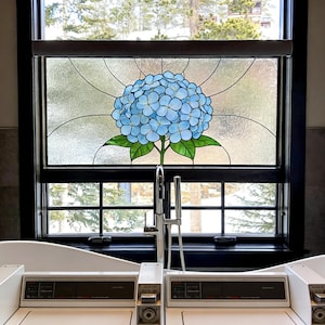 May include: A stained glass window panel featuring a central blue hydrangea flower with green leaves, set within a dark wooden frame. Below the window, two white washing machines are positioned side-by-side with a silver faucet in between.
