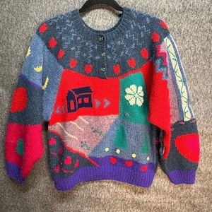 Könnte beinhalten: Ein farbenfroher Patchwork-Pullover mit rundem Ausschnitt und Knopfverschluss. Der Pullover zeigt eine Vielzahl geometrischer Formen und Muster in Rot, Blau, Grün und Lila. Ein Haus und eine Blume gehören zu den Designs.