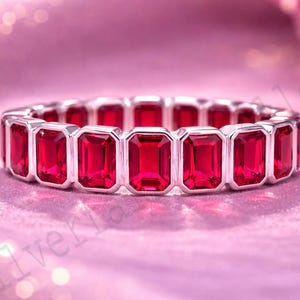 May include: A silver ring with a continuous band of rectangular, vibrant red gemstones. The ring is set against a soft pink background, with a blurred bokeh effect adding depth. The gemstones are evenly spaced.
