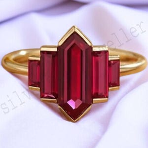 May include: A gold ring featuring a central, elongated, deep red gemstone flanked by two smaller rectangular red stones on each side. The ring has a geometric design and is set against a white fabric background.