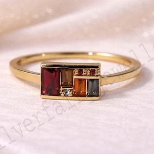 May include: A gold ring with a rectangular setting, showcasing a mosaic of coloured gemstones. The gemstones include red, brown, orange, and blue hues. The ring has a slender band and a minimalist design.