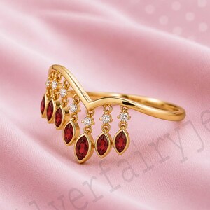 May include: A gold V-shaped ring featuring a series of dangling charms, each set with a marquise-cut deep red gemstone. Small, round, clear stones are set at the top of each charm, creating a delicate fringe effect along the band's curve.