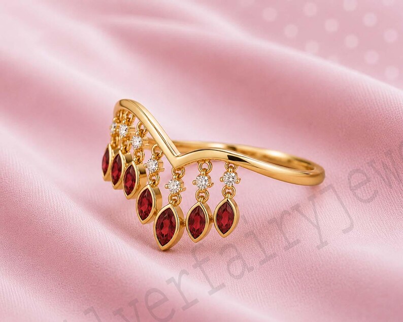 May include: A gold V-shaped ring featuring a series of dangling charms, each set with a marquise-cut deep red gemstone. Small, round, clear stones are set at the top of each charm, creating a delicate fringe effect along the band's curve.