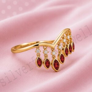 May include: A gold V-shaped ring featuring a series of dangling charms. Each charm consists of a small round clear stone set above a marquise-cut red gemstone. The ring has a polished gold band that follows a chevron silhouette along the finger.