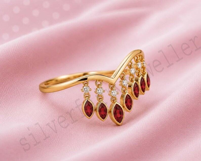 May include: A gold V-shaped ring featuring a series of dangling charms. Each charm consists of a small round clear stone set above a marquise-cut red gemstone. The ring has a polished gold band that follows a chevron silhouette along the finger.