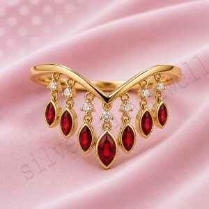 May include: A gold V-shaped ring featuring a series of dangling charms. Each charm consists of a small round clear stone set above a marquise-cut red gemstone. The ring has a polished gold band that follows a chevron silhouette along the finger.