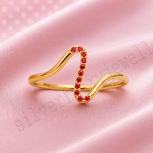 May include: A gold ring featuring a wavy, serpentine band design. The center section of the ring is accented with a vertical row of small, round, vibrant red gemstones set into the polished gold surface, creating a distinct and continuous curve.