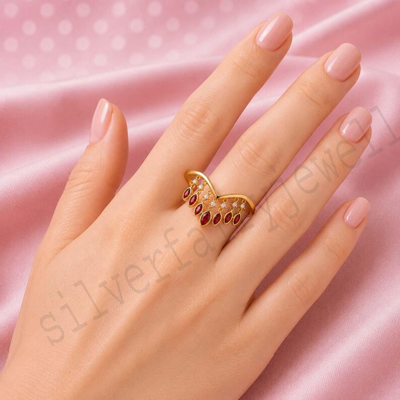 May include: A gold V-shaped ring featuring a series of dangling marquise-cut red gemstones and small round white accent stones. The ring has a polished gold band that follows a chevron silhouette, with the red stones suspended from the metal frame.
