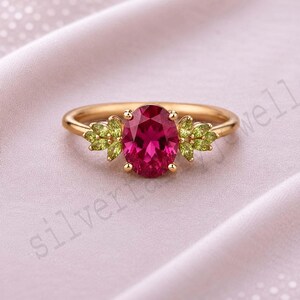 Può includere: Un anello d'oro con una grande pietra preziosa rosso rubino di taglio ovale come punto focale. Ai lati della pietra centrale, grappoli di pietre preziose verde chiaro più piccole. La fascia dell'anello è in oro lucido. L'anello è su uno sfondo rosato tenue.