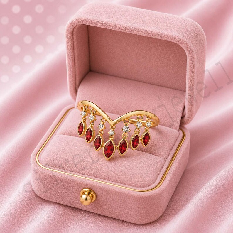 May include: A gold chevron ring featuring a series of dangling marquise-cut red stones and small round clear stones. The ring is displayed inside an open, square, pink velvet jewellery box with a gold-toned clasp on the front edge.