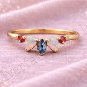 Puede incluir: Un anillo de oro con un diseño curvo, con una gema azul central en forma de marquesa flanqueada por dos ópalos en forma de pera y dos pequeñas gemas rojas. El anillo está sobre un fondo rosa suave.